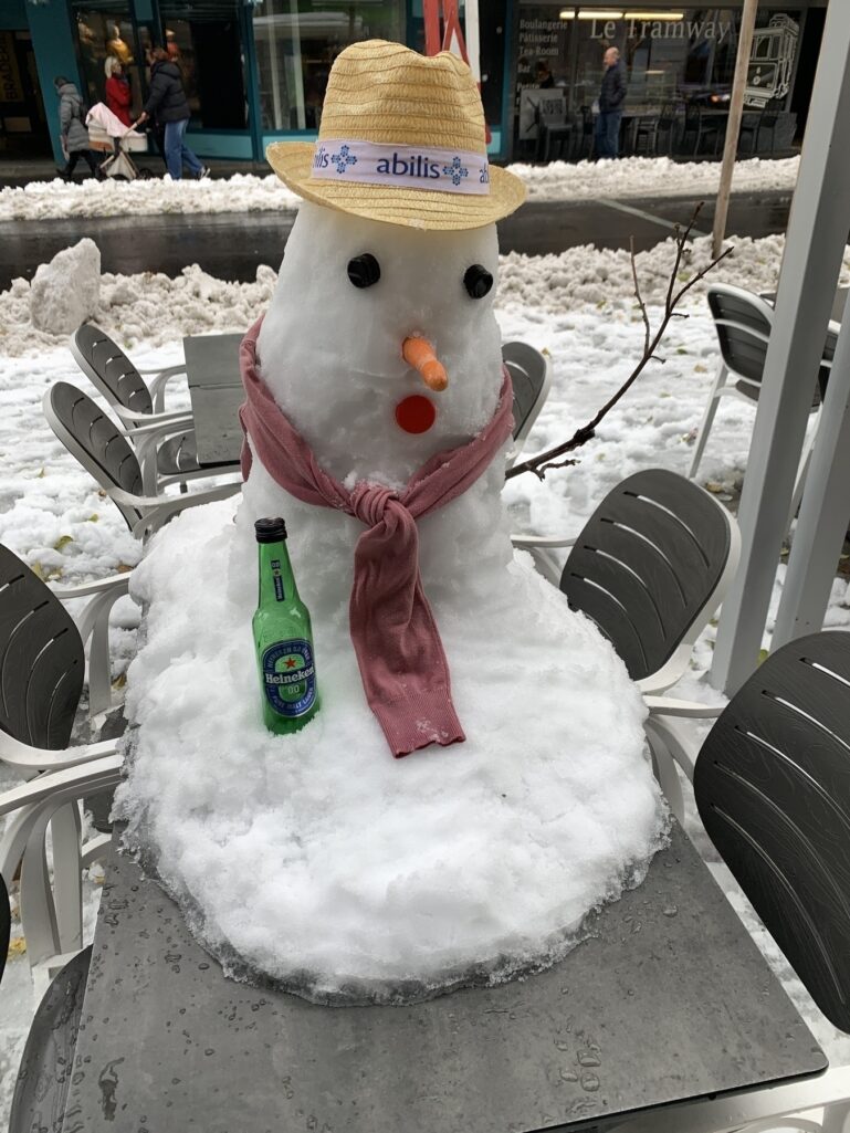 Un bonhomme de neige portant un chapeau de paille et une écharpe est assis sur une table avec une bouteille à côté dans une rue enneigée.