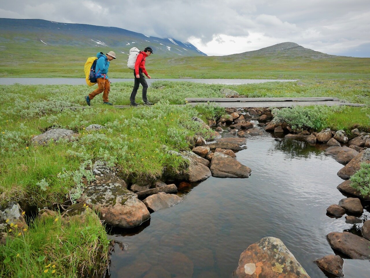 Une personne en orange avec un sac à dos blanc et une autre en bleu avec un sac à dos jaune, vues de côté, marchent et s'apprêtent à franchir une petite passerelle sur un petit bras bras de lac entouré de verdure sous un ciel menaçant. 
One person in orange with a white backpack and another in blue with a yellow backpack, seen from the side, are walking and about to cross a small footbridge over a small arm of the lake surrounded by greenery under a threatening sky.