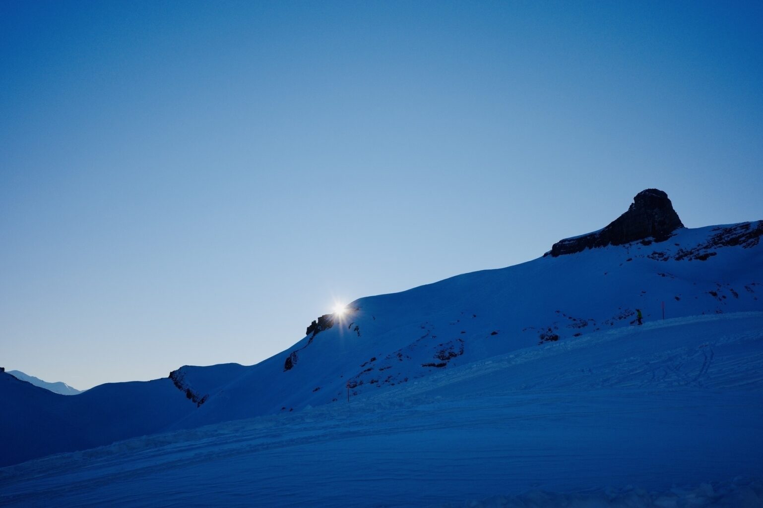 Soleil disparaissant derrière une arête montagneuse enneigée. 