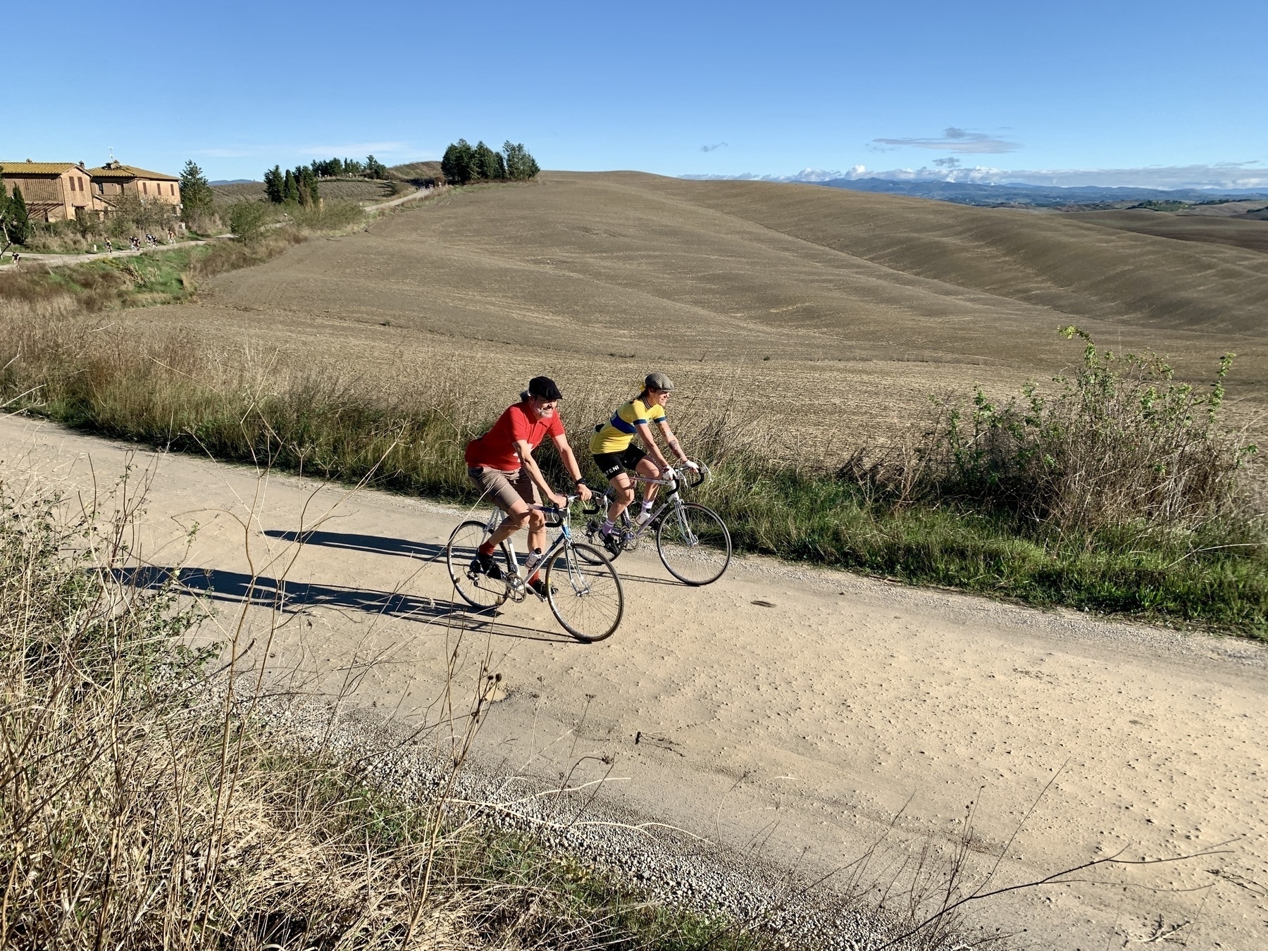 Deux cyclistes roulent sur un chemin de terre dans un paysage rural avec de la végétation verte et une maison au loin.