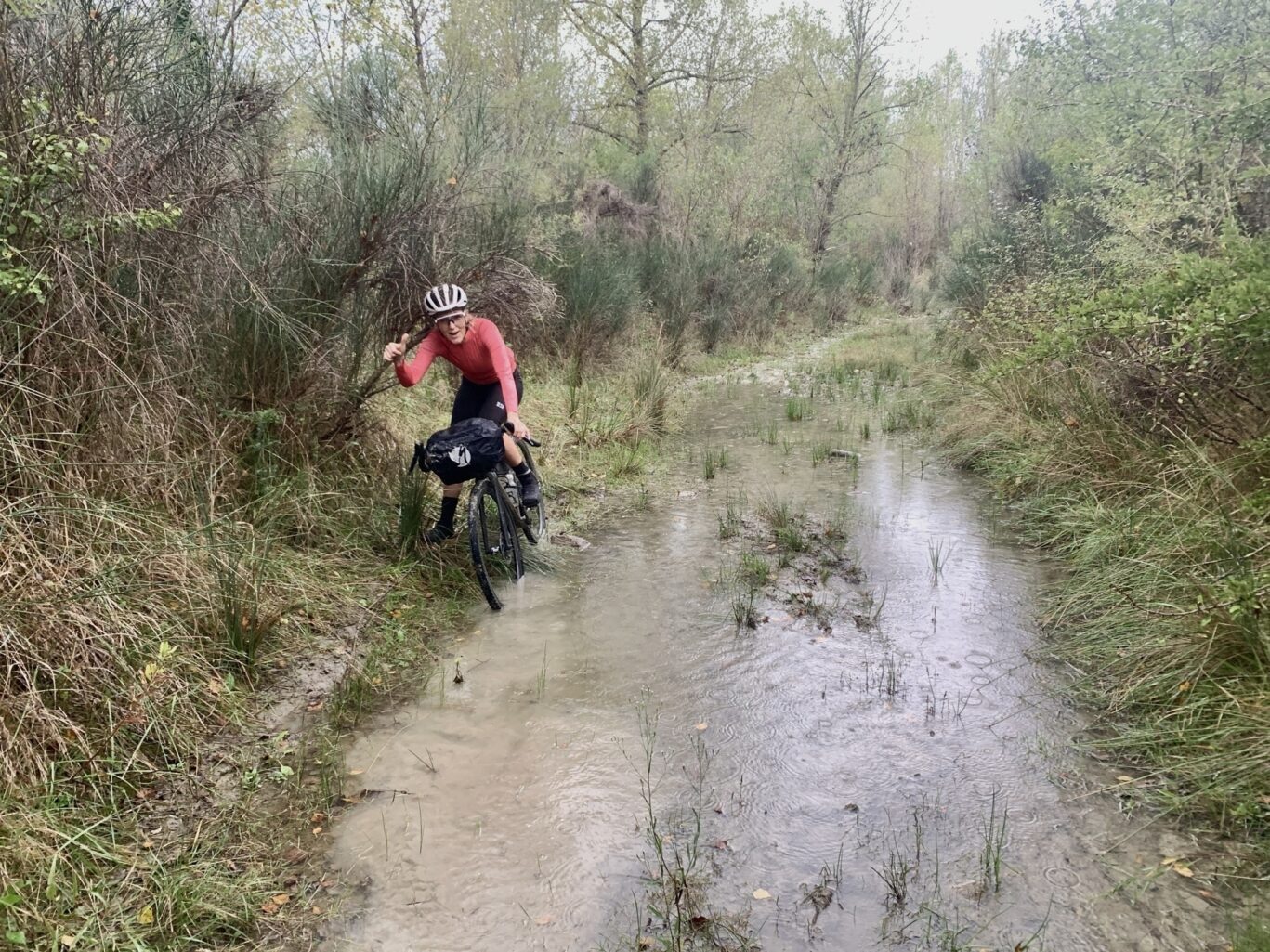 Une cycliste emprunte un sentier boueux et gorgé d'eau, entouré d'une végétation dense.