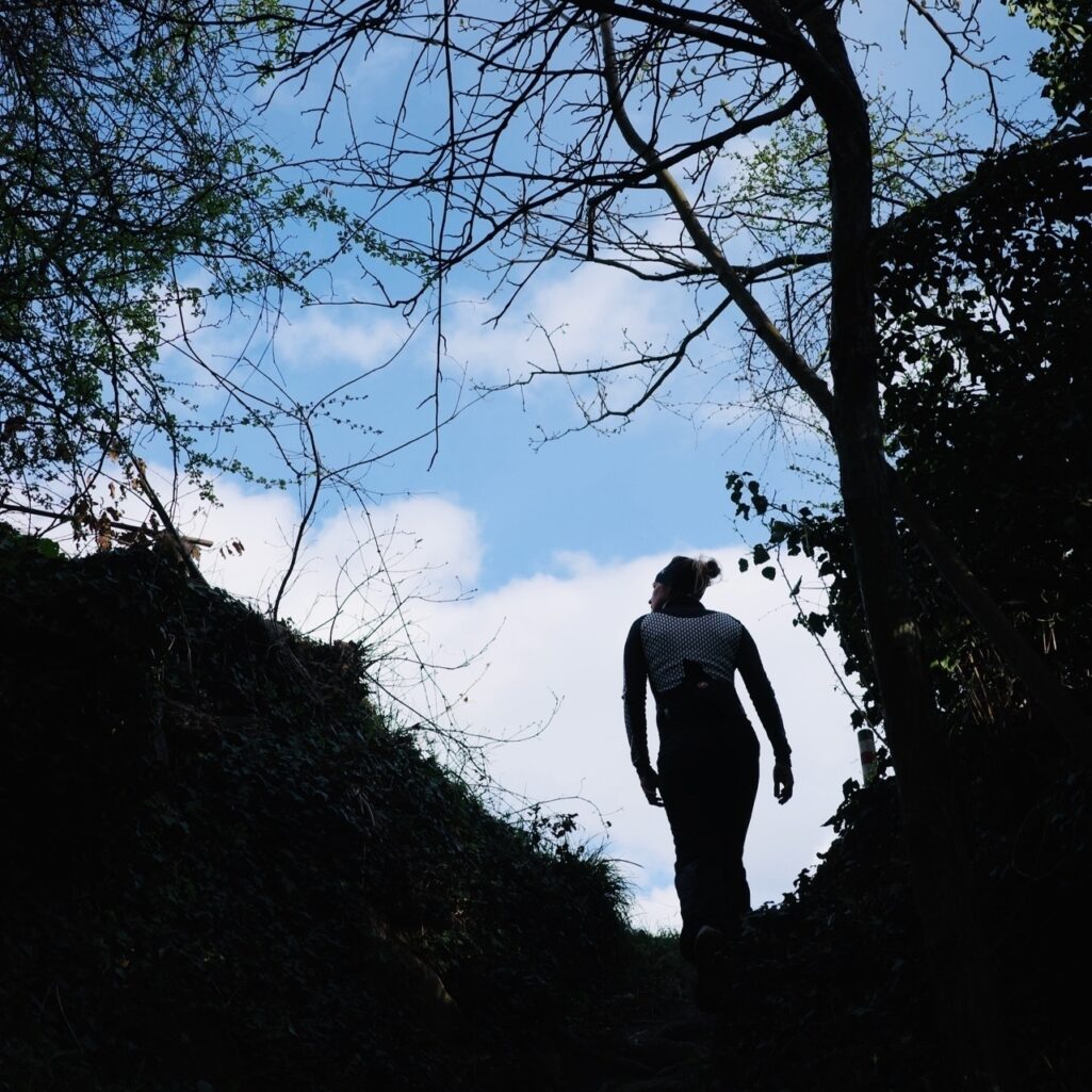 Image carrée, marcheuse en silhouette et branches d’arbre en contrejour et contreplongée sur fond de ciel bleu avec quelques nuages.