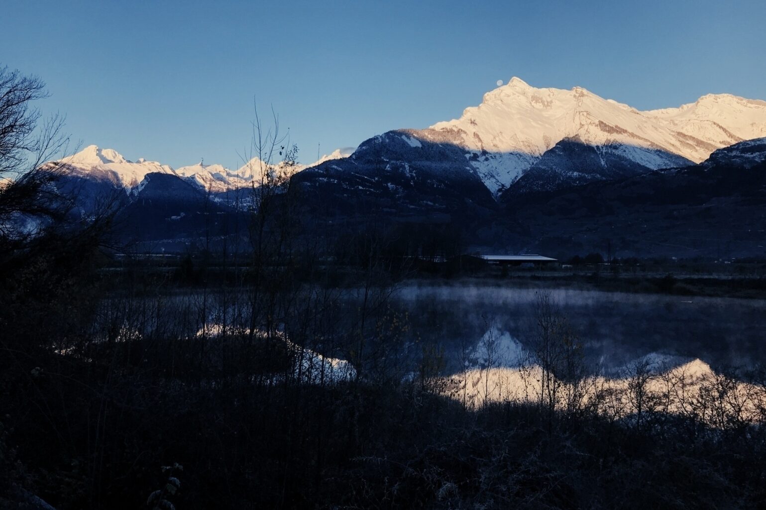 Montagne enneigée au lever du soleil, avec la lune qui s'arrête à disparaître derrière l'arête, avec reflet dans un étang au premier plan.