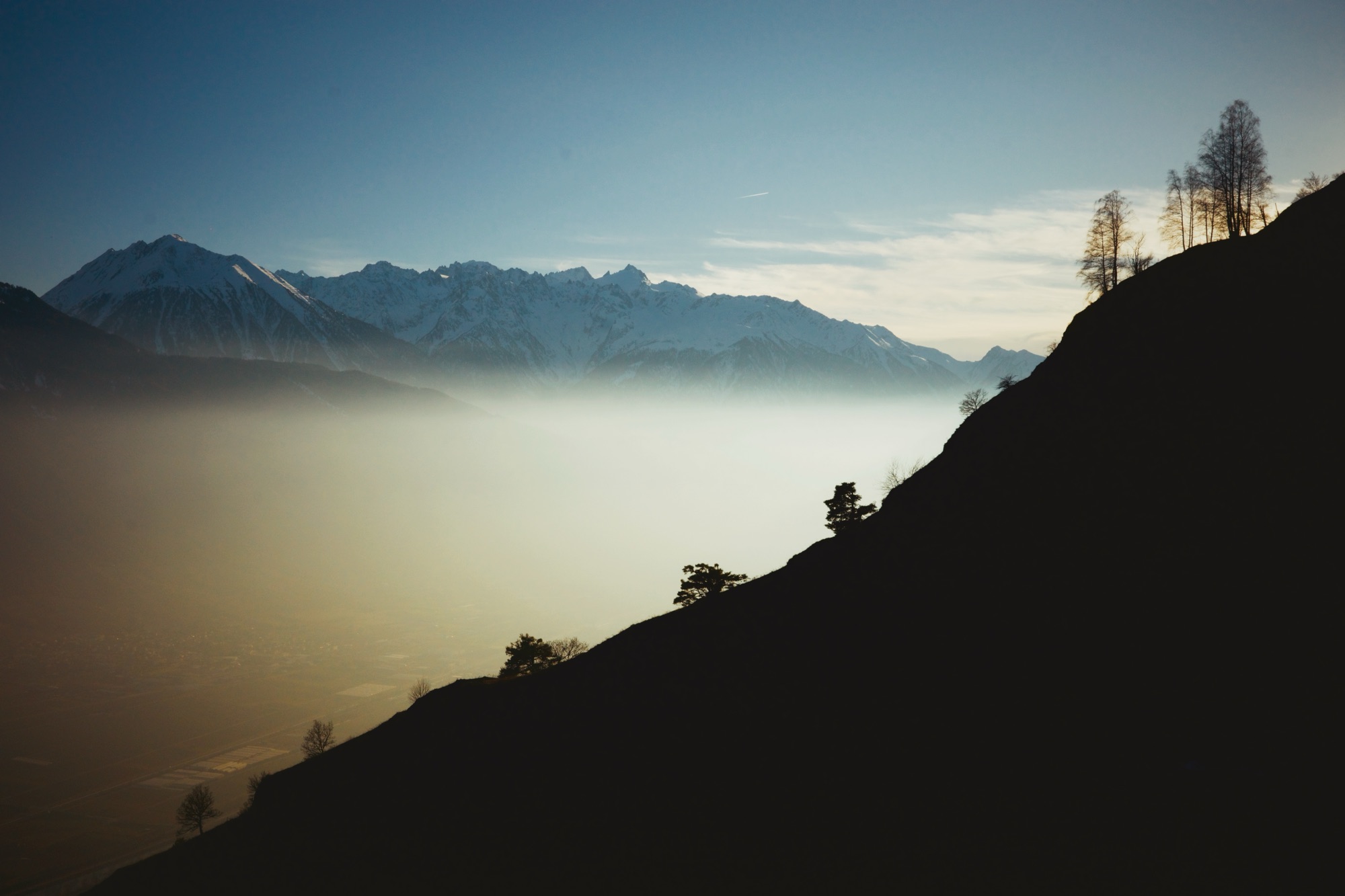 Montagnes enneigées sur une vallée brumeuse, vues depuis une colline sombre.