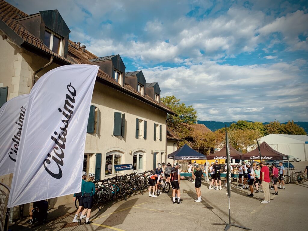 Les cyclistes se rassemblent devant un charmant bâtiment au toit sombre et aux murs blancs. Les vélos sont alignés et les gens s'affairent sous des tentes noires siglées Ciclismo. Une grande bannière Ciclismo est déployée bien en évidence.