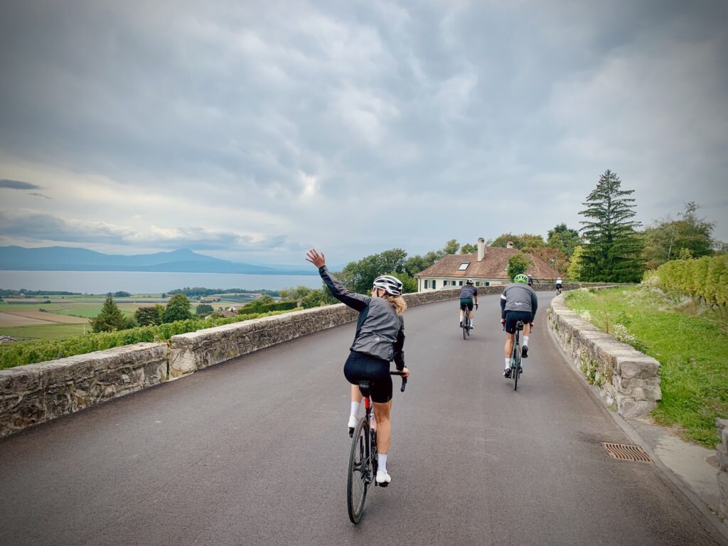 Trois cyclistes profitent d'une balade pittoresque sur une route sinueuse surplombant un lac, le Léman, sur la gauche, avec des montagnes dans le fond. L'une d'eux salue, profitant du magnifique panorama de la campagne suisse.