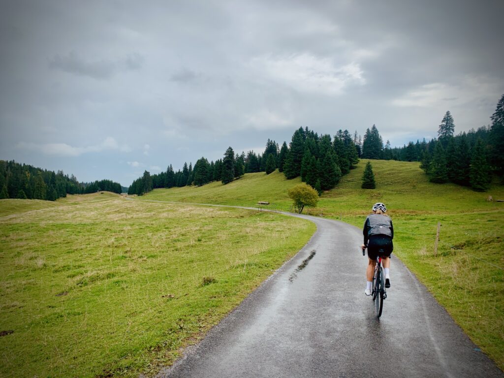 Une cycliste vêtu d'une veste sombre roule sur une route asphaltée pittoresque et sinueuse. La route traverse des collines verdoyantes et une forêt de conifères sous un ciel nuageux. L'ambiance générale est sereine et paisible.