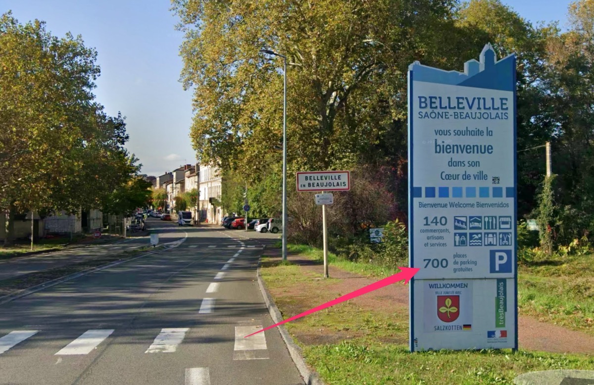 La route à l'entrée de Belleville-en-Beaujolais. Des arbres entourent la chaussée à gauche et à droite à l'arrière-plan. Devant, à la hauteur d'un passage piéton dont il cache en partie l'accès, un panneau indique que la ville compte 700 place de parc gratuites et 140 commerces.