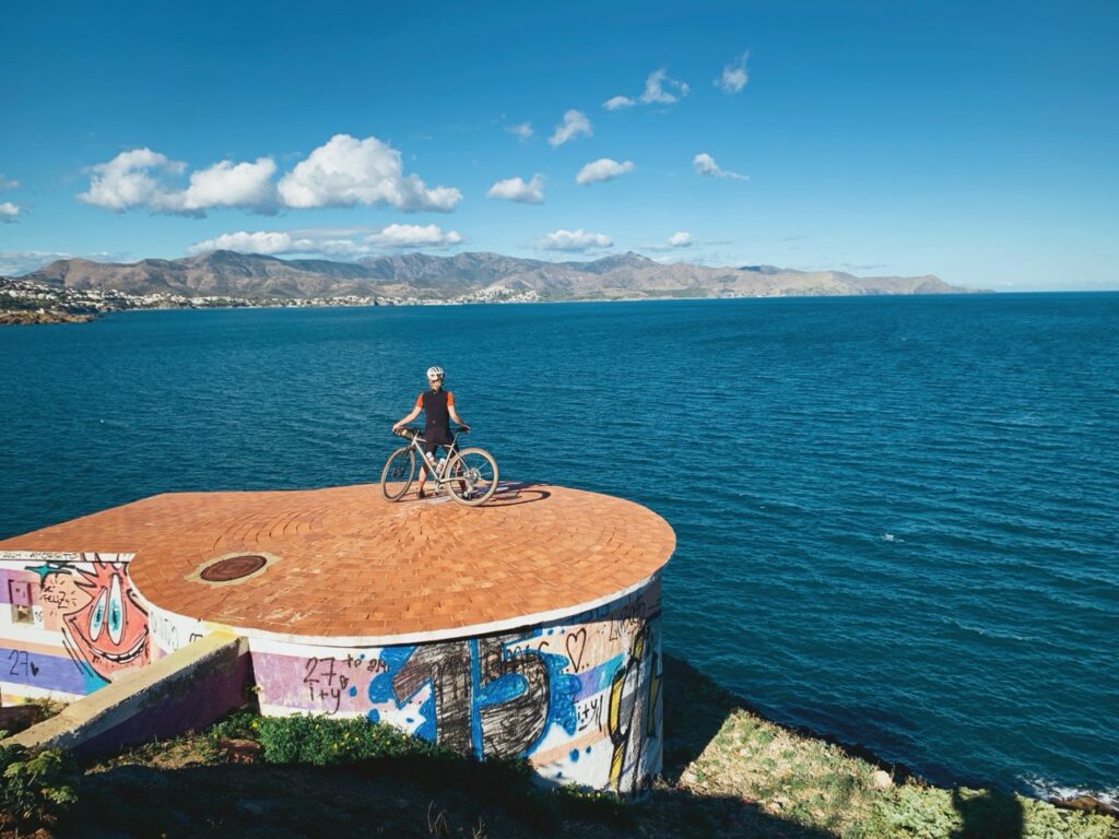Cycliste sur une plate-forme ornée de graffitis colorés, en bord de mer, avec vue sur les montagnes lointaines et un ciel bleu dégagé.