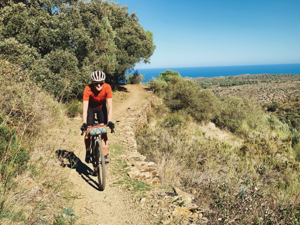 Cycliste souriante parcourant un sentier de montagne avec vue sur la mer sous un ciel bleu en journée ensoleillée.