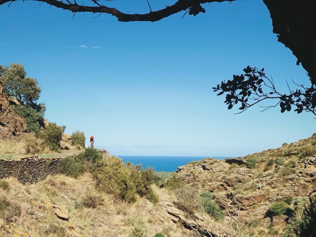 Paysage pittoresque de montagne avec vue sur la mer sous un ciel bleu clair entouré de végétation naturelle et rochers. Une cycliste se détache au loin.