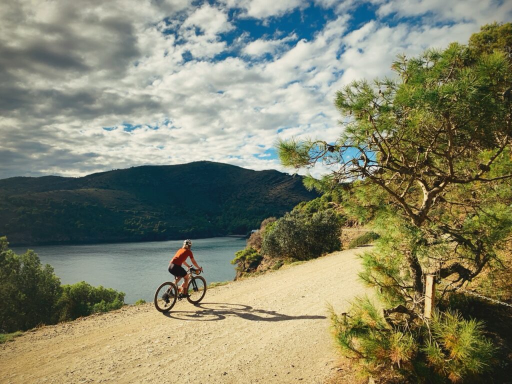 Cycliste sur un chemin de terre longeant la mer, avec des collines et des arbres sous un ciel partiellement nuageux.