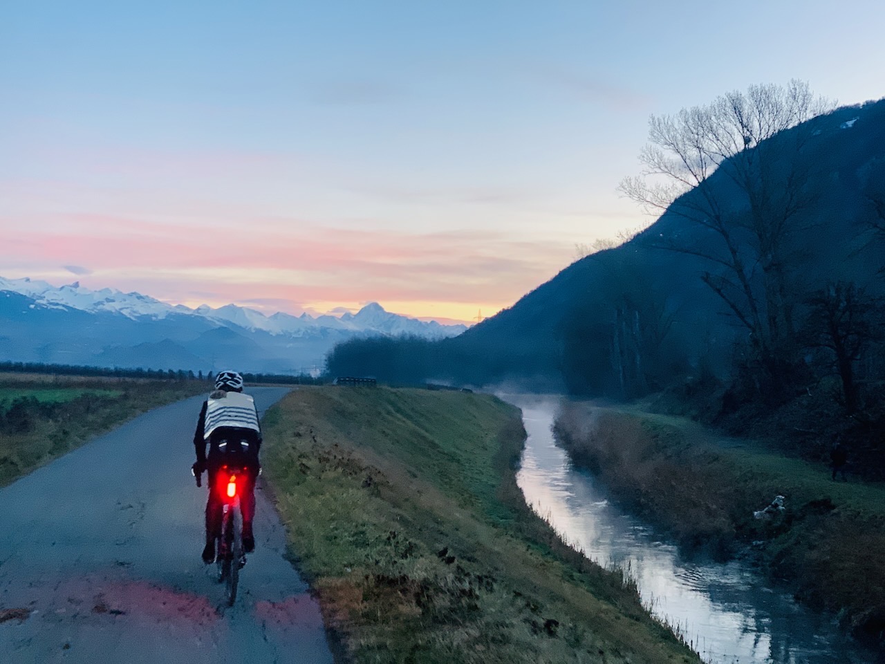 Cycliste vue de dos avec son feu arrière rouge sur une route longeant un petit canal à droite. La scène est au lever du jour, avec un ciel légèrement orange sur des montagnes à l’horizon.