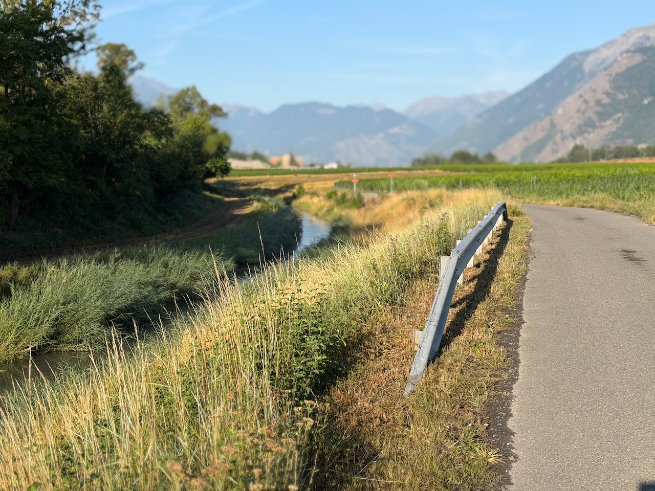 Scène rurale paisible, avec un chemin sinueux s'évanouissant au loin au milieu d'une végétation luxuriante bordant un canal sur la gauche et de montagnes lointaines.