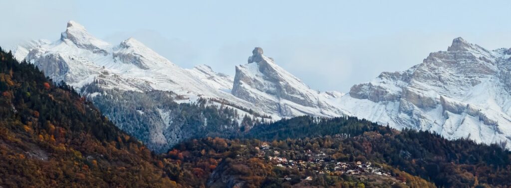 Vue panoramique spectaculaire des Alpes. Les pentes sont drapées de forêts d'automne vibrantes, menant à un petit village blotti au creux du paysage. L'arrière-plan est dominé par une chaîne de pics enneigés et imposants, capturant la beauté brute des montagnes en saison de transition.